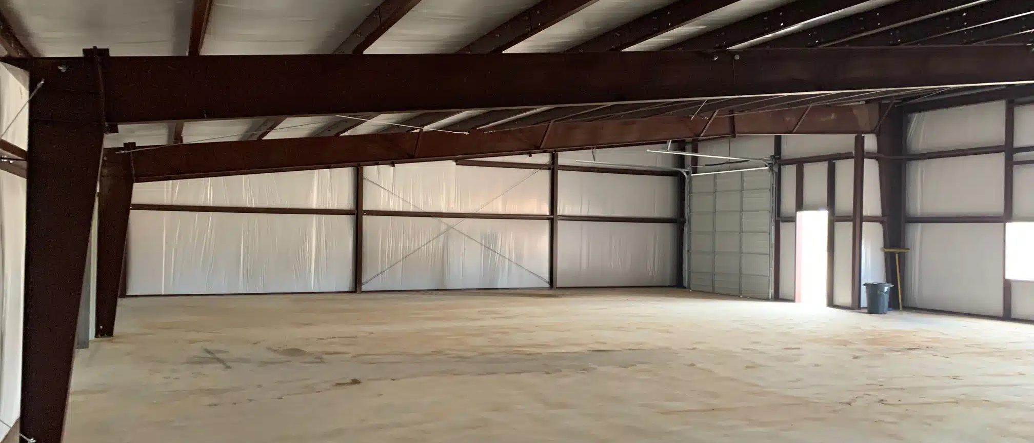 Inside a red iron steel building: rafters, insulated white walls, a concrete floor and an overhead door