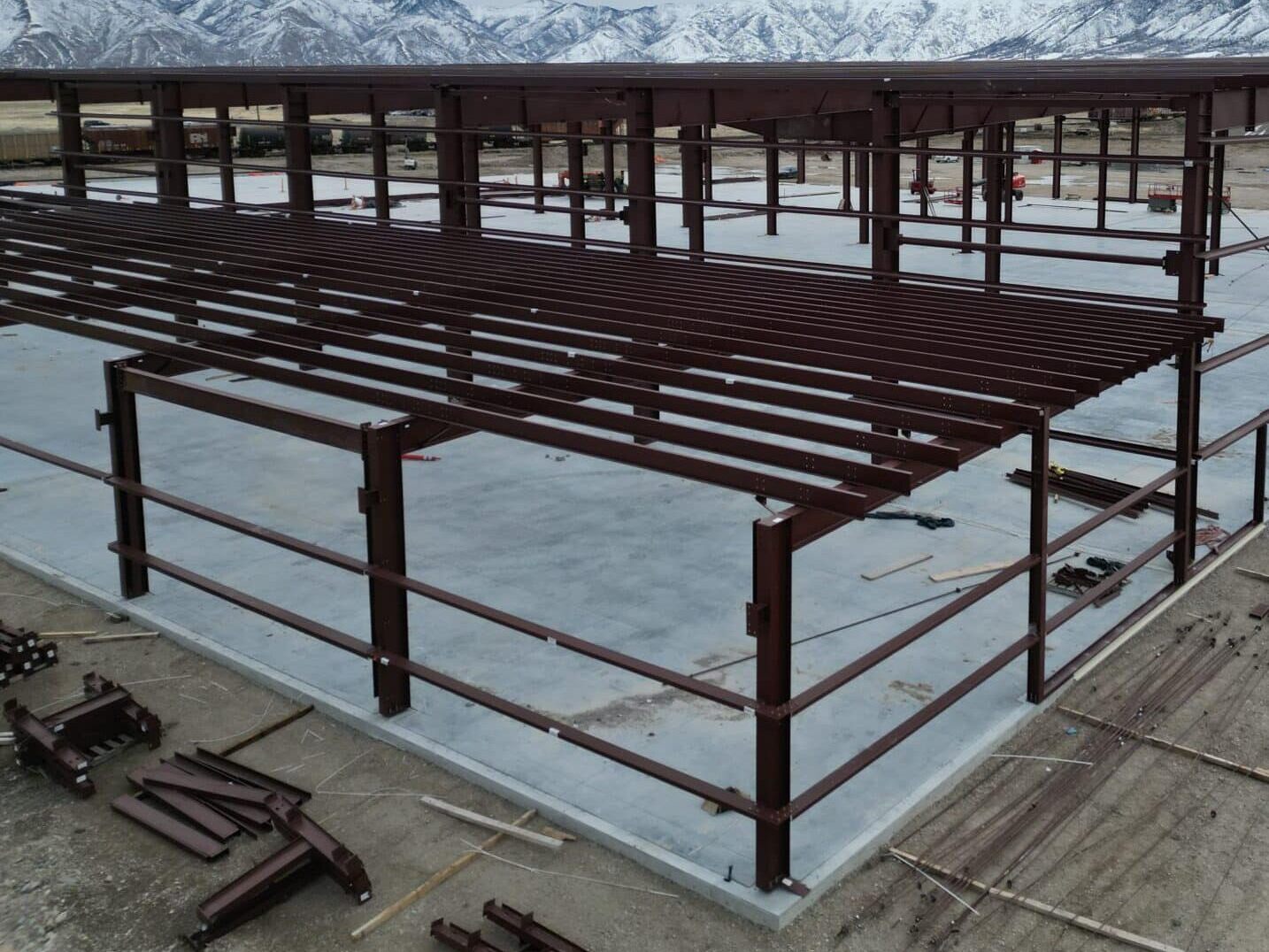 Red iron columns, beams and purlins of a large steel building going up on a concrete slab, with snowy mountains behind